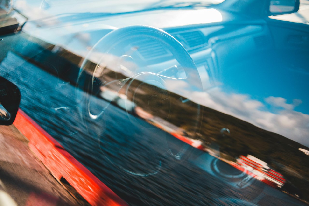 Reflexionen eines Lenkrads und blauen Himmels auf der Windschutzscheibe eines roten Autos.